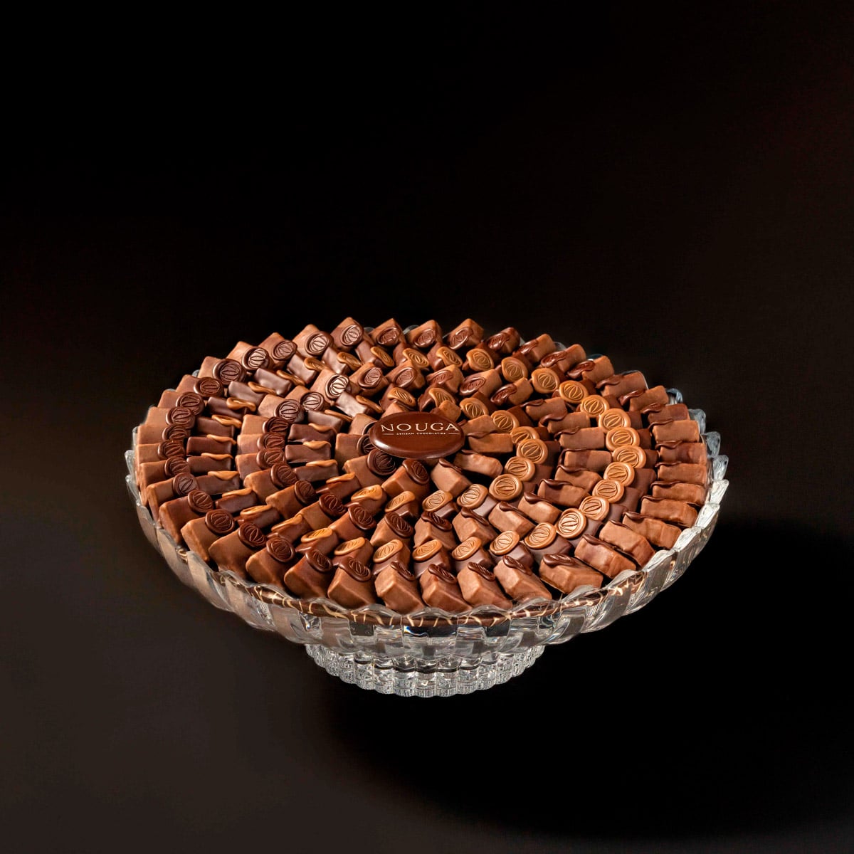 Stack of chocolate bonbons on a clear glass stand with 'Nouga' branding against a dark background
