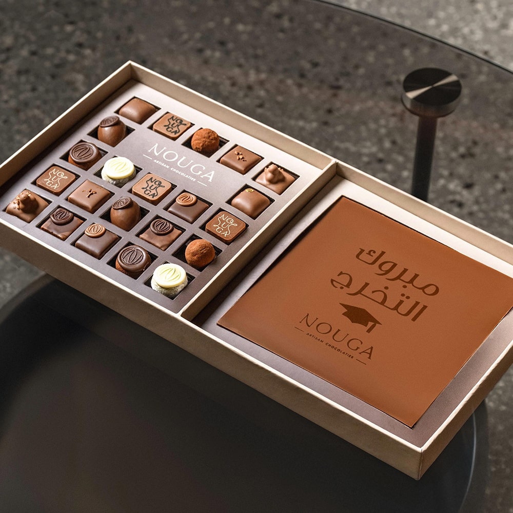 Open box of Nouga chocolates with a brown chocolate message slab on a dark surface