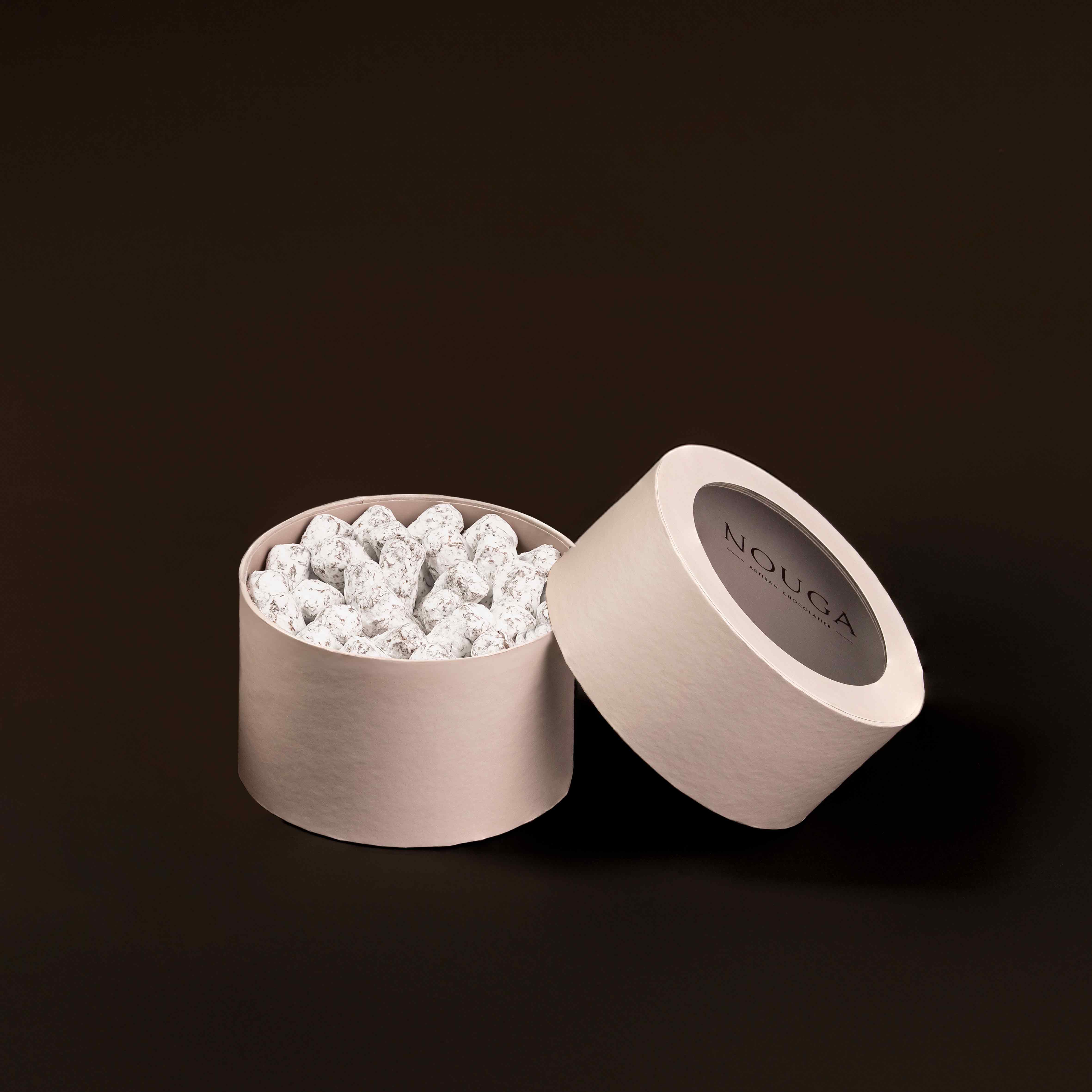 Open beige container with white chocolate barks on a dark background, featuring the brand 'NOUGA'.