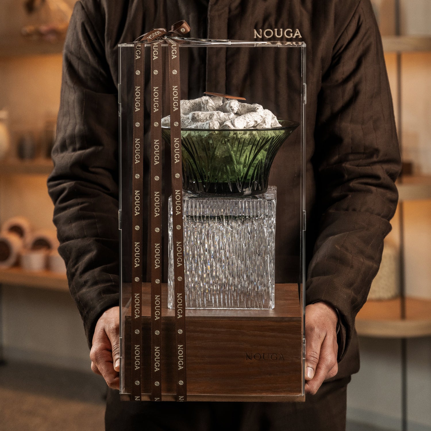 Person holding a transparent packaging container with a crystal tray filled with chocolate, wood base is branded 'Nouga'. Gift adorned with ribbons.