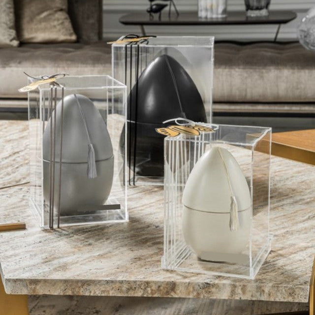 Decorative egg objects in various sizes placed inside clear acrylic boxes on a marble surface with a blurred background