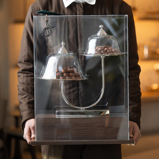 Person holding a transparent display case with two glass domes containing chocolate truffles.