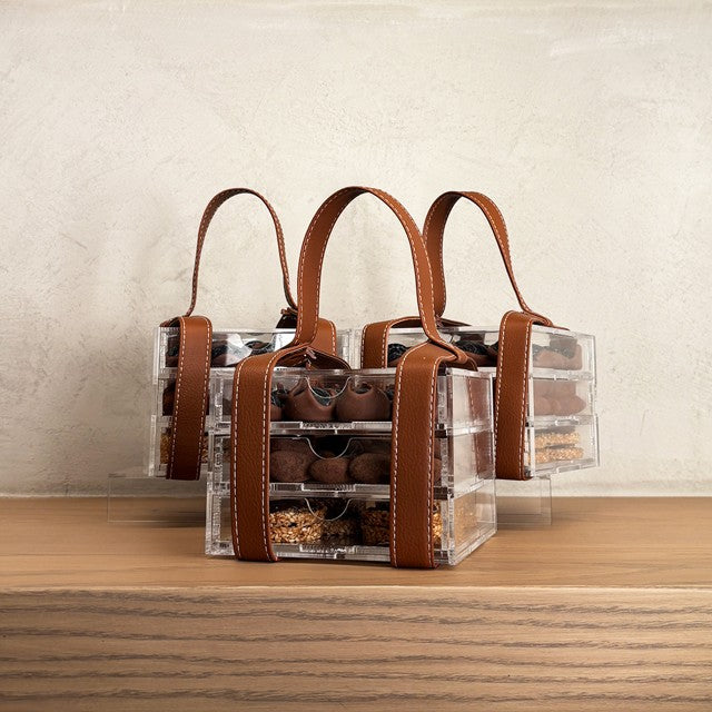 Clear acrylic chocolate storage containers with brown leather handles on a wooden surface.