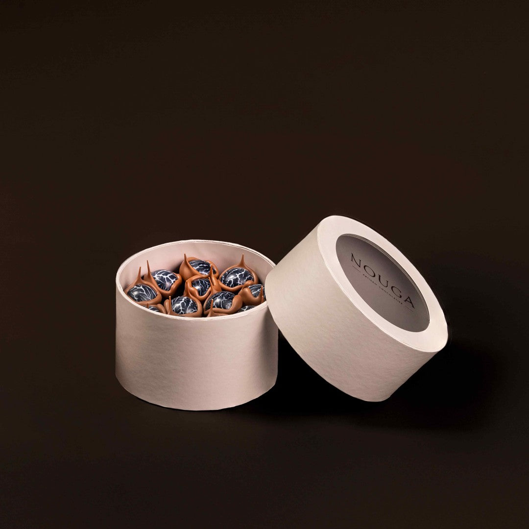 Open beige container with decorative bonbons on a dark background, featuring the brand 'NOUGA'.
