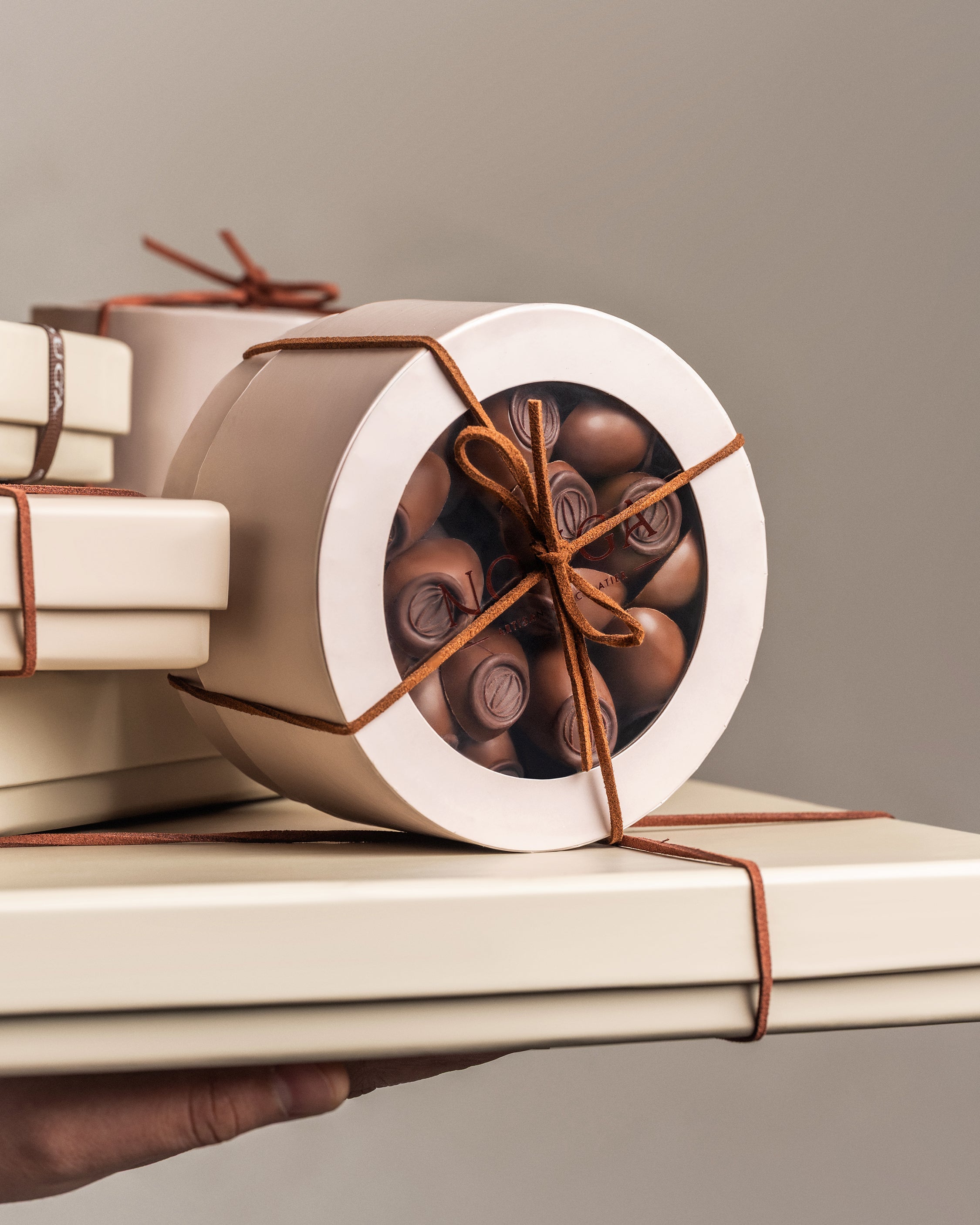 Circle box filled with chocolate, wrapped with leather string and top lid shows Nouga brand logo