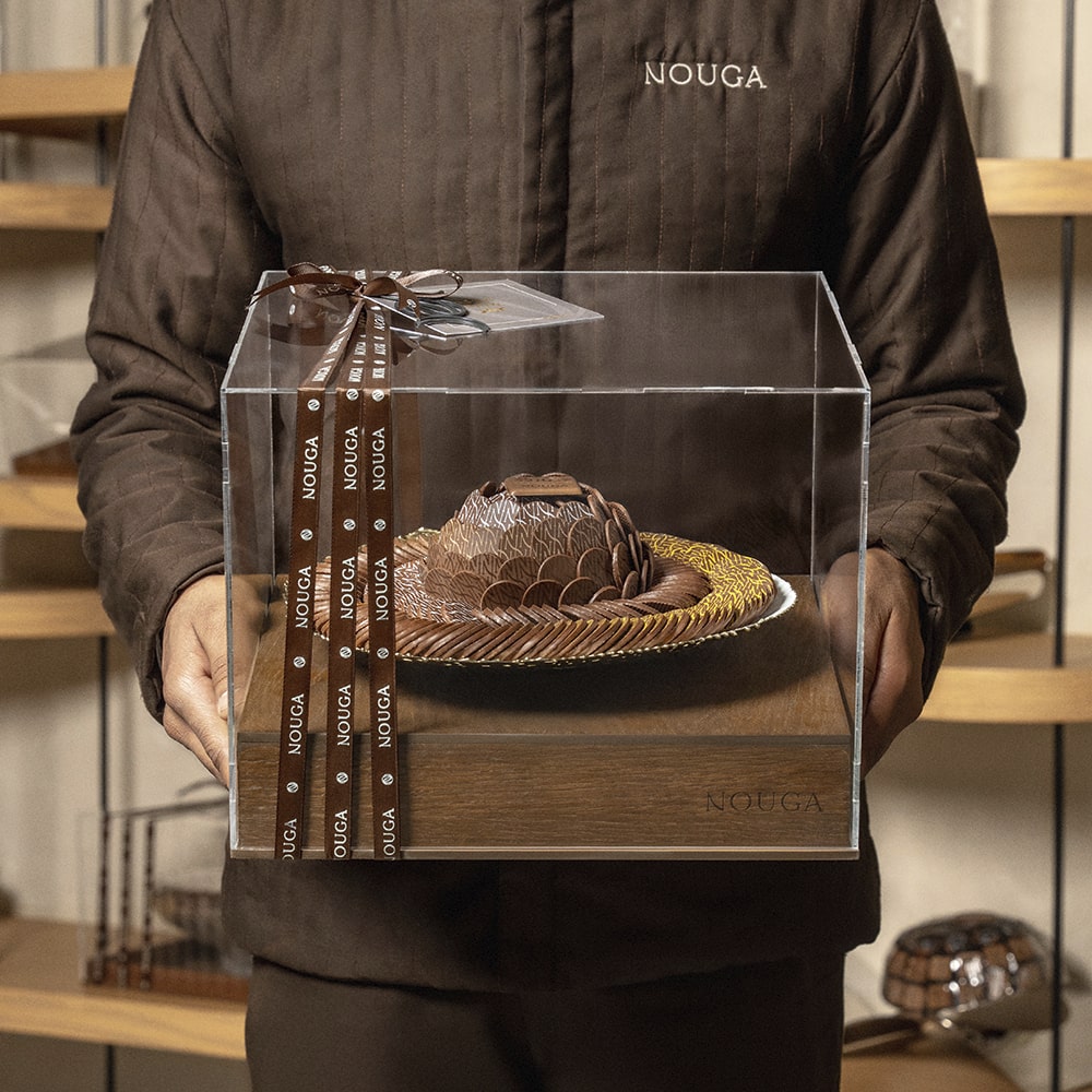 Person holding a chocolate tray with sculpture in a clear box with 'Nouga' branding.