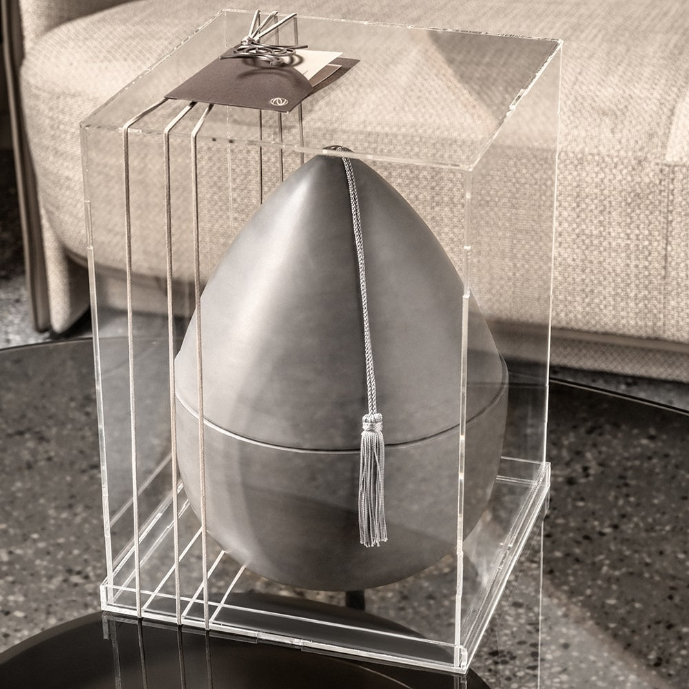 Decorative gray egg ceramic tray with a tassel in a clear box, placed on a reflective glass surface.