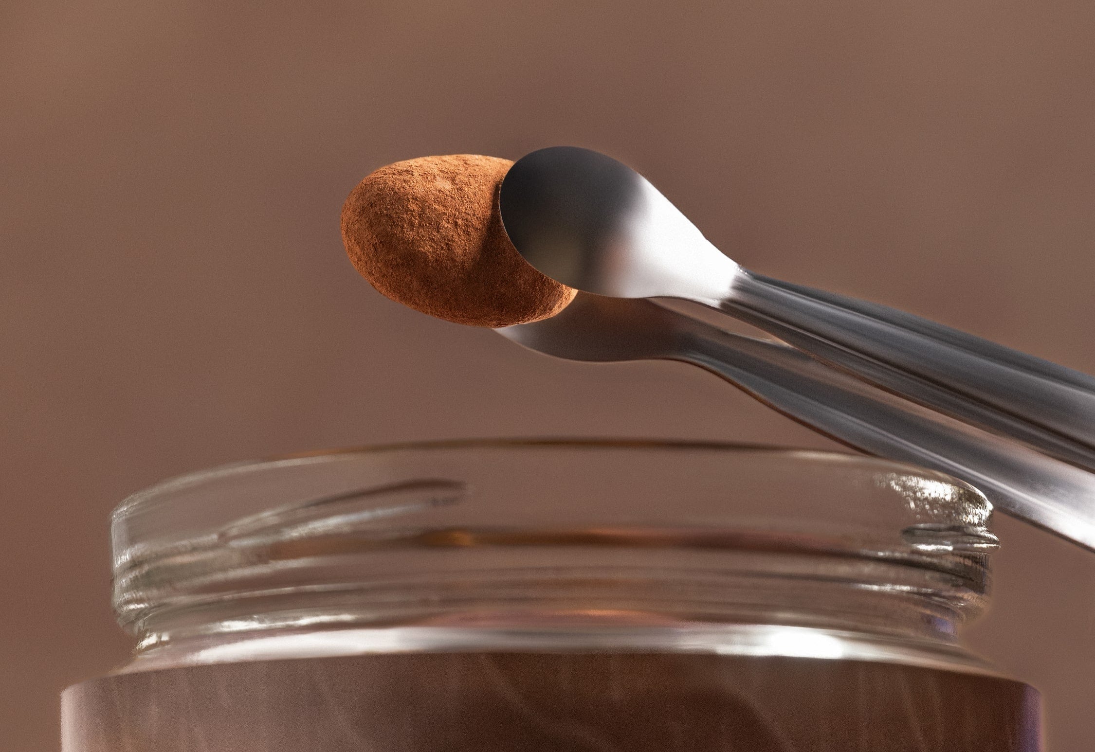 Tweezers holding brown cocoa dusted almond dragees above an open jar on a brown background