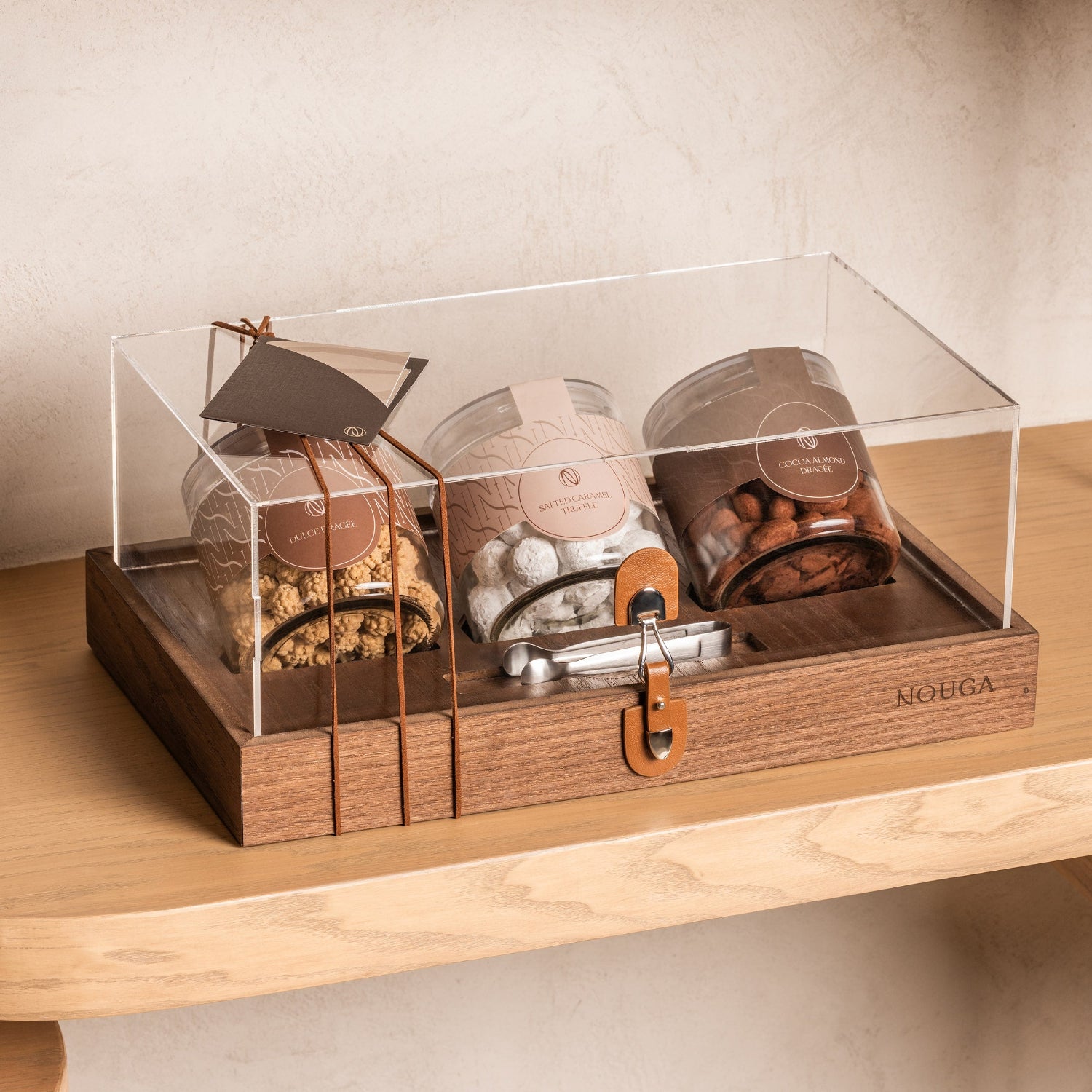 Wooden gift box with transparent lid containing three wrapped jars on a wooden surface filled with chocolate. The box includes tweezers.