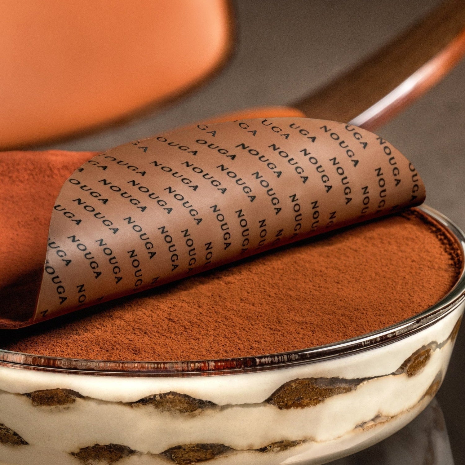 Classic italian tiramisu with layers of ladyfingers and marscapone creme, topped with tempered chocolate wave that is branded with the word 'Nouga'.