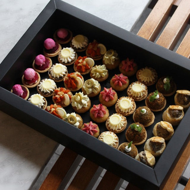 Assorted small pastries and canapés in a black box on a wooden and marble surface.