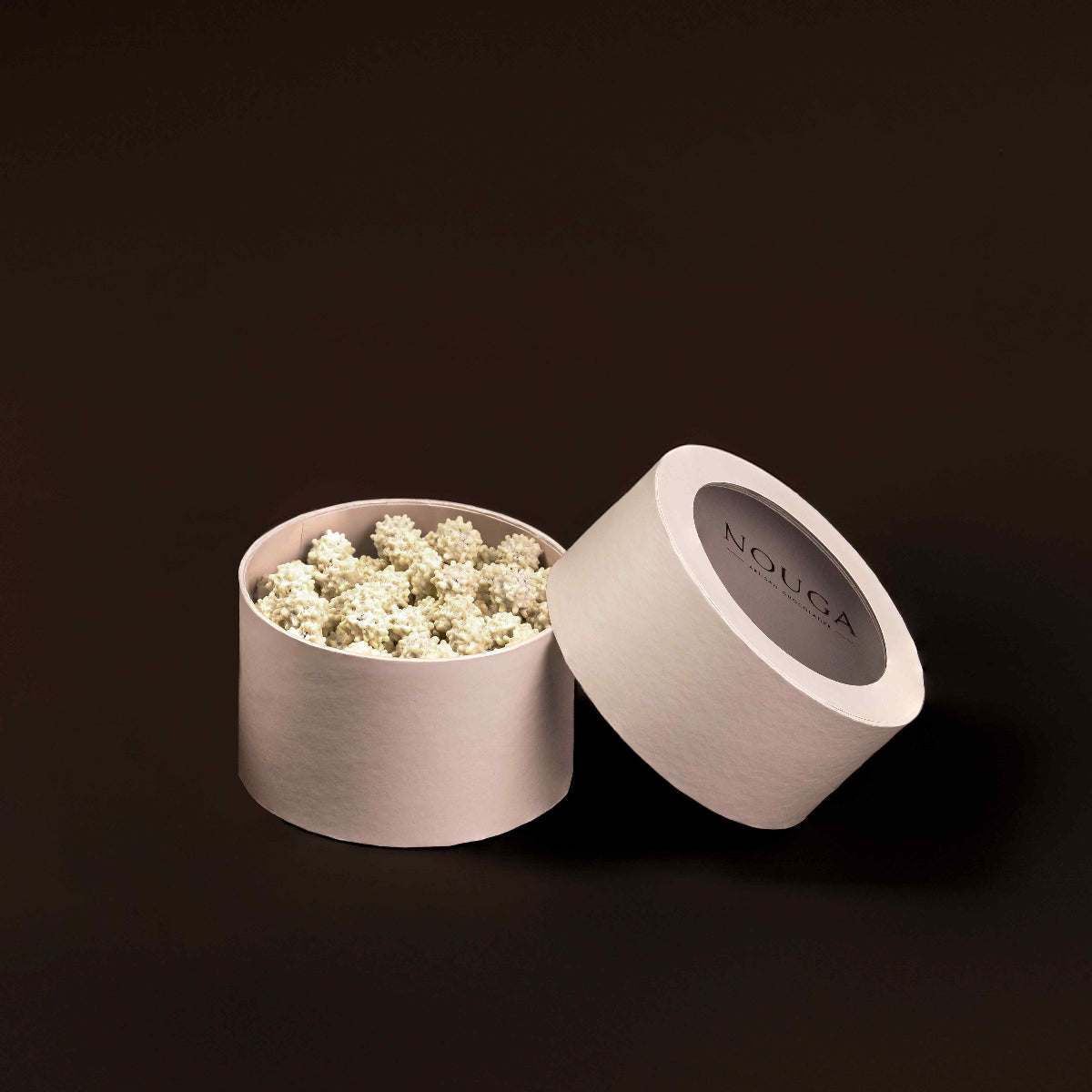 Open beige circle chocolate box with white yuzu dragee on a dark brown background. Lid featuring the brand 'NOUGA'.